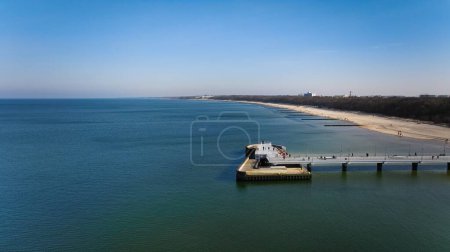 Muelle de Kolobrzeg en una soleada tarde de febrero. Clima sin viento, mar tranquilo sin una sola ola, turistas paseando por el muelle. Atmósfera serena capturada en una foto en stock.