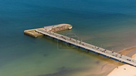 Kolobrzeg pier on a sunny February afternoon. Windless weather, calm sea without a single wave, tourists strolling on the pier. Serene atmosphere captured in a stock photo.