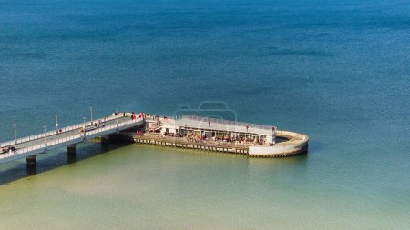 Kolobrzeg pier on a sunny February afternoon. Windless weather, calm sea without a single wave, tourists strolling on the pier. Serene atmosphere captured in a stock photo.