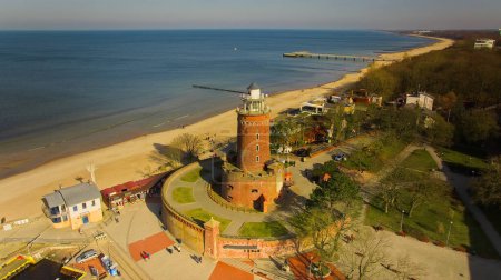 February in Kolobrzeg, Poland: A serene scene featuring a red brick lighthouse, people enjoying a sunny day on the sandy beach, and a tranquil sea without any waves.