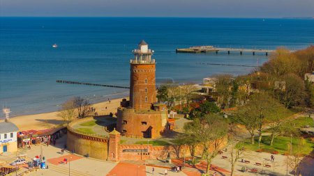 February in Kolobrzeg, Poland: A serene scene featuring a red brick lighthouse, people enjoying a sunny day on the sandy beach, and a tranquil sea without any waves.