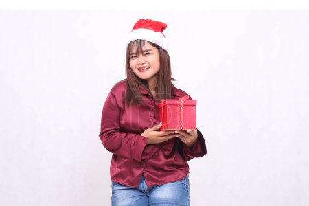 Photo for Photo portrait of beautiful cheerful Asian girl in her 20s carrying boxed gifts in Christmas Santa Claus hat wearing shiny modern red shirt seen on white background for promotion and advertising - Royalty Free Image