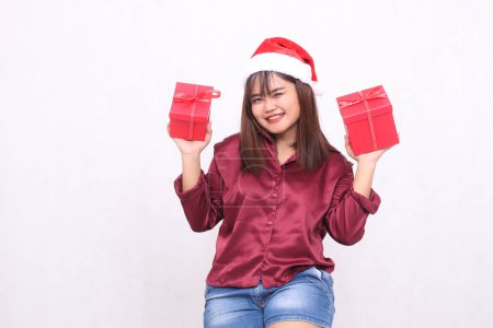 Photo for Beautiful young asian indonesian girl carrying gift box at christmas santa claus hat modern shiny red shirt outfit looking at camera on white background for promotion and advertising - Royalty Free Image
