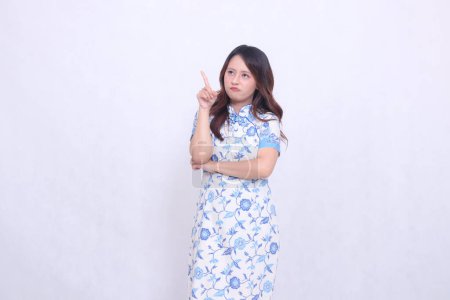 Beautiful Chinese Javanese woman smiling thoughtfully wearing a modern Chinese dress with her hand pointing obliquely to the right on a white background. happy Chinese New Year
