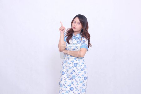 Beautiful Javanese Chinese girl pouting wearing a modern Chinese dress with her arms crossed pointing at the top left on a white background. happy Chinese New Year