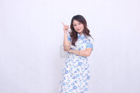 Beautiful Chinese Javanese woman smiling wearing a modern Chinese dress with her arms crossed pointing at the top left on a white background. happy Chinese New Year