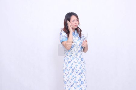 Photo for Beautiful Indonesian Chinese woman looking obliquely at top left wearing modern Chinese blue dress holding tablet while making phone call isolated on white background - Royalty Free Image