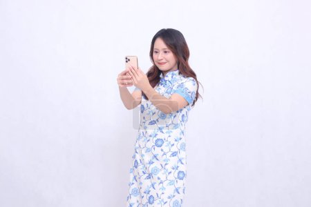 Photo for Young asian javanese chinese woman smile wearing blue chinese dress looking at cellphone and operating it isolated white background - Royalty Free Image