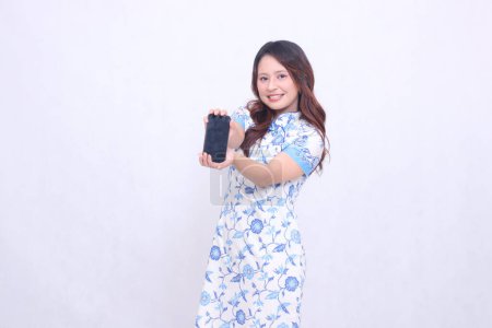 Photo for Young asian javanese chinese girl wearing chinese traditional blue dress cheerful promoting and holding gadget on mobile phone screen fore isolated white background - Royalty Free Image