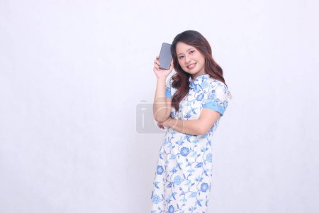 Photo for Young Indonesian Javanese woman wearing a blue traditional Chinese dress cheerful with her arms holding a gadget showing an upright cellphone screen isolated on a white background - Royalty Free Image
