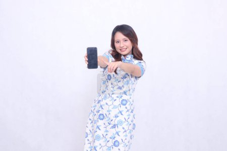 Photo for Young Indonesian Javanese woman wearing blue traditional Chinese dress happy with arms wide holding gadget showing cellphone screen forward pointing isolated white background - Royalty Free Image