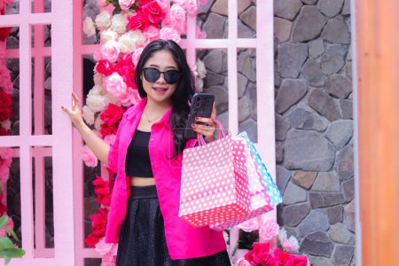 Photo for Beautiful Asian girl in pink shirt smiling checking orders via cellphone gadget, standing carrying paperbag and credit card at outdoor cafe for promotion of fashion, lifestyle, catalog - Royalty Free Image