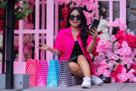 Photo for Beautiful asian woman in pink shirt wearing glasses smiling on video call showing her shopping, sitting between paperbags in outdoor cafe for promotion of fashion, lifestyle, catalog - Royalty Free Image