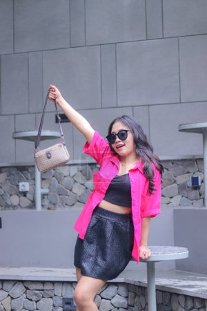 Photo for Sexy indonesia career woman in pink shirt happy glasses hands holding bag up leaning on table for fashion, lifestyle, banner, promo, business content - Royalty Free Image