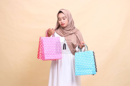 the charm of a young Muslim woman wearing a hijab, sullenly looking at and carrying the contents of a colorful paper bag from top-down shopping. for advertising, lifestyle, banners and Ramadan
