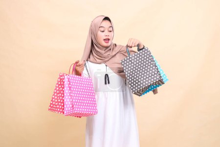 the charm of a young Asian Muslim woman wearing a hijab is surprised to the left looking at and carrying the contents of a colorful paper bag from shopping from top to bottom