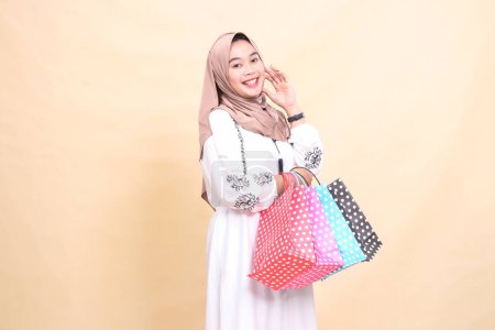 Photo for The story of a young Asian Muslim woman wearing a hijab cheerfully holds her cheek and carries a shopping bag of various variations up and down. for advertising, culture, Eid and Ramadan - Royalty Free Image