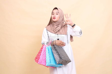 the story of a young Asian Muslim girl wearing a hijab with an expression of kissing an okay gesture and carrying shopping bags. for advertising, culture, Eid and Ramadan