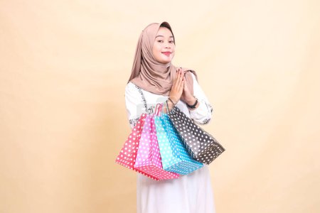 Photo for The story of a young Asian Muslim woman wearing a hijab with a smile, a gesture of welcome (sawadikap) and carrying shopping bags. for advertising, culture, Eid and Ramadan - Royalty Free Image