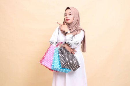 die geschichte einer jungen indonesischen muslimischen frau, die einen hijab trägt, der nach rechts zeigt und eine paperbag shopping bag trägt. für Werbung, Kultur, Eid und Ramadan