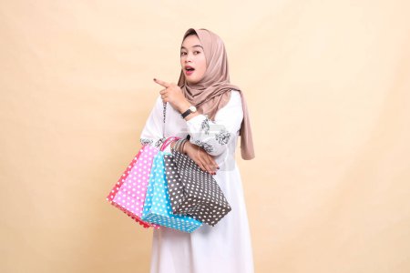 The story of a young Asian Muslim woman wearing a hijab was shocked, pointing to the right and carrying a paper bag. for advertising, culture, Eid and Ramadan