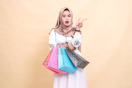 Photo for A young Asian Muslim girl wearing a hijab was shocked, pointing to the left and carrying a paper bag. for advertising, culture, Eid and Ramadan - Royalty Free Image