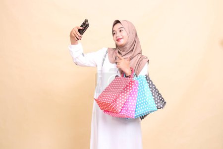 a young Asian Muslim woman wearing a hijab takes a selfie pointing at a cellphone camera and carrying a paperbag shopping bag. for advertising, culture, Eid and Ramadan
