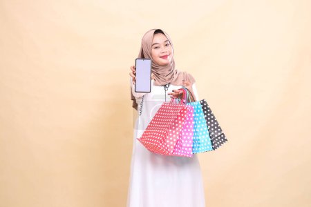 una joven indonesia musulmana con un hiyab sonríe con gracia, muestra una pantalla de teléfono celular y lleva una bolsa de papel. para publicidad, tecnología, Eid y Ramadán