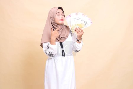 portrait of a beautiful indonesia Muslim girl wearing a hijab, smiling, holding and pointing at a gift or fitrah to the left on Eid day. used for advertising, giveaways, Eid and Ramadan