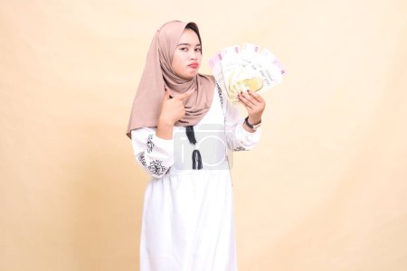Portrait of a beautiful Asian Muslim woman wearing a hijab, pouting gracefully, holding and pointing to a gift or fitrah to the left on Eid day. used for advertising, giveaways, Eid and Ramadan