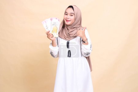 retrato de una hermosa mujer musulmana asiática con un hiyab sonriendo con un gesto de pulgares hacia arriba sosteniendo un regalo o fitrah a la derecha en el día del Eid. utilizado para la publicidad, regalos, Eid y Ramadán