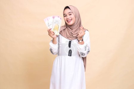 portrait of a beautiful Asian Muslim woman wearing a hijab smiling cheerfully as a gesture of love (saranghaeyo) holding a gift or fitrah to the right on Eid day. used for advertising, Eid and Ramadan