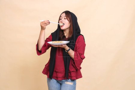 Photo for Beautiful mature indonesia woman wearing a red hijab poses, bites into dimsum with chopsticks up and carries a plate containing dimsum (Chinese food). used for food, health and culinary content - Royalty Free Image