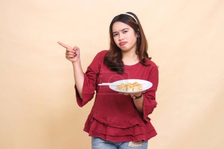 Photo for Beautiful young Asian girl in red shirt is disappointed carrying a plate containing dimsum (Chinese food) and chopsticks while pointing to the right. used for food, health and lifestyle content - Royalty Free Image