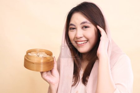 Photo for Cheerful indonesia woman wearing a hijab at right angles, holding her hair back and holding a wooden bowl filled with chopsticks and delicious dimsum - Royalty Free Image