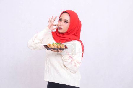 Photo for Indonesia woman wearing a hijab is cheerful while gesturing okay, good, delicious, kissing and carrying a plate containing sushi (Japanese food). Beautiful Muslim women are used for advertising - Royalty Free Image