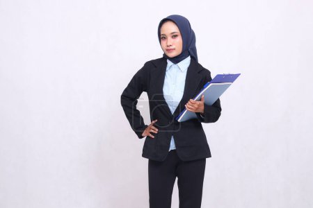 Une femme de bureau indonésienne exclusive portant un hijab se tient élégamment avec ses mains sur sa taille et porte un stylo, un presse-papiers et un cahier. Belle femme musulmane en chemise bleue