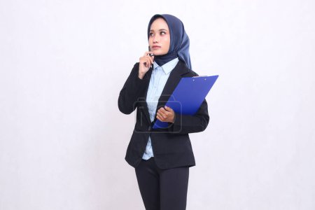 Asiatique mature bureau femme en hijab debout pensant à la droite portant un stylo et étreignant un presse-papiers. Belles femmes musulmanes portant des chemises bleues pour les entreprises, les finances et les entreprises