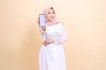 Photo for Girl asian muslim wearing a hijab, smiling cheerfully, holding a cellphone gadget in her hand in front of her with her arms crossed. Lifestyle, business and finance concept - Royalty Free Image