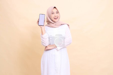 Photo for Girl asian muslim wearing a hijab smiling happily holding a cell phone gadget in her hand with her arms crossed. Lifestyle, business and finance concept - Royalty Free Image