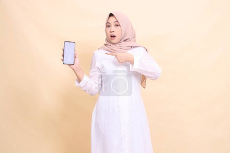 Photo for Young Muslim woman asian wearing a hijab is shocked holding her cellphone gadget to the right in her hand while pointing. Lifestyle, business and commercial concept - Royalty Free Image