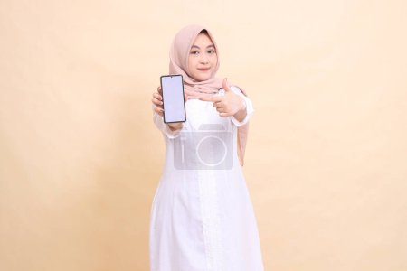 Photo for Asian Muslim woman wearing a hijab smiling, holding a cell phone gadget in her hand in front of her while pointing at it. Lifestyle, business and finance concepta - Royalty Free Image