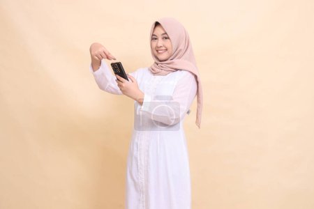 Photo for Woman asian muslim wearing a hijab smiles cheerfully at the camera, operating a cellphone gadget with both hands while pointing. Lifestyle, technology and promotion concept - Royalty Free Image