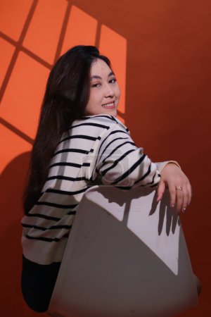 A beautiful cheerful asian woman holds and sits on a chair in a dark studio, the light shines on her forming a window pattern. Portrait of woman in projection light (spotlight) in studio