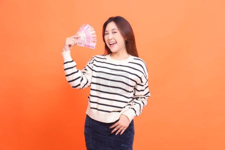 an Asian girl with a cheerful expression at the camera stands while holding several rupiah notes and holding her skirt. for fashion, business and finance concept