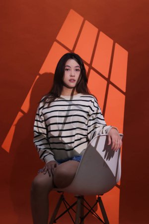 A beautiful asian girl in a dark studio, light shines on her through the curtains, forming a window pattern. Portrait of woman in projection light (spotlight) in studio