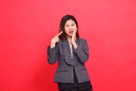 der Gesichtsausdruck einer asiatischen Büroangestellten, die Schmerzen mit Zahnschmerzen hat, während sie in einer grauen Jacke und schwarzem Minirock auf rotem Hintergrund auf sie zeigt. für Gesundheits-, Geschäfts- und Werbekonzepte