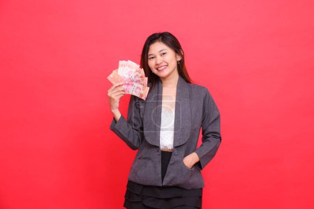 Ausdruck einer fröhlichen indonesischen Bürofrau, die in grauer Jacke und rotem Rock die EC-Karte und das Geld aufrecht und in der Tasche in die Kamera hält. für Transaktion, Geschäft und Werbung