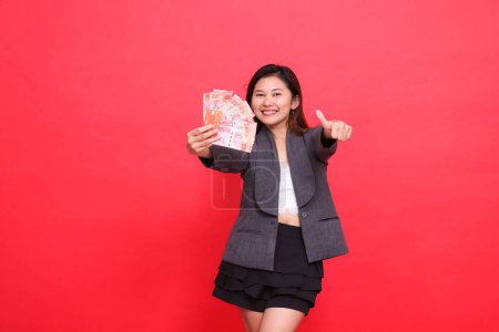 geste d'une femme de carrière indonésienne heureuse tenant une carte de crédit et de l'argent devant un panneau pouce levé portant une veste et une jupe sur un fond rouge. pour transaction, affaires et publicité