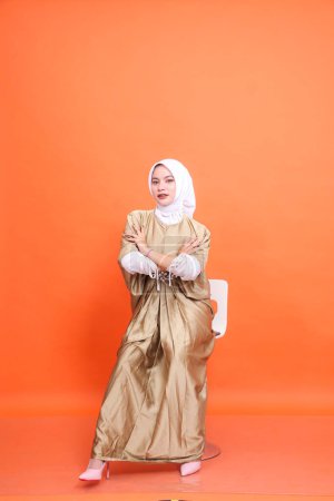 Full length studio portrait of young asian muslim woman with arms crossed with white hijab in wearing kaftan, high heels sitting on chair isolated on orange background. Concept of work, fashion